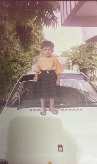 Standing on the hood of a car as a child, wearing a yellow top and dark skirt.