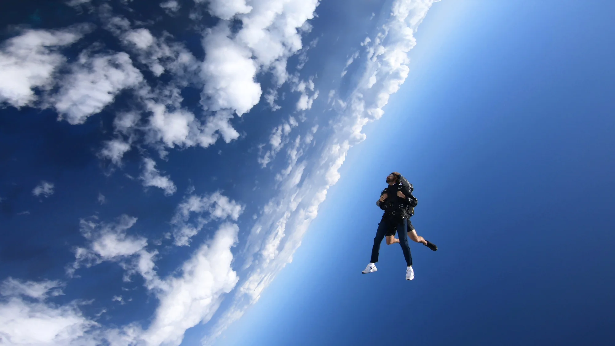 Tandem skydiving against an open blue sky and a diagonal bank of clouds.