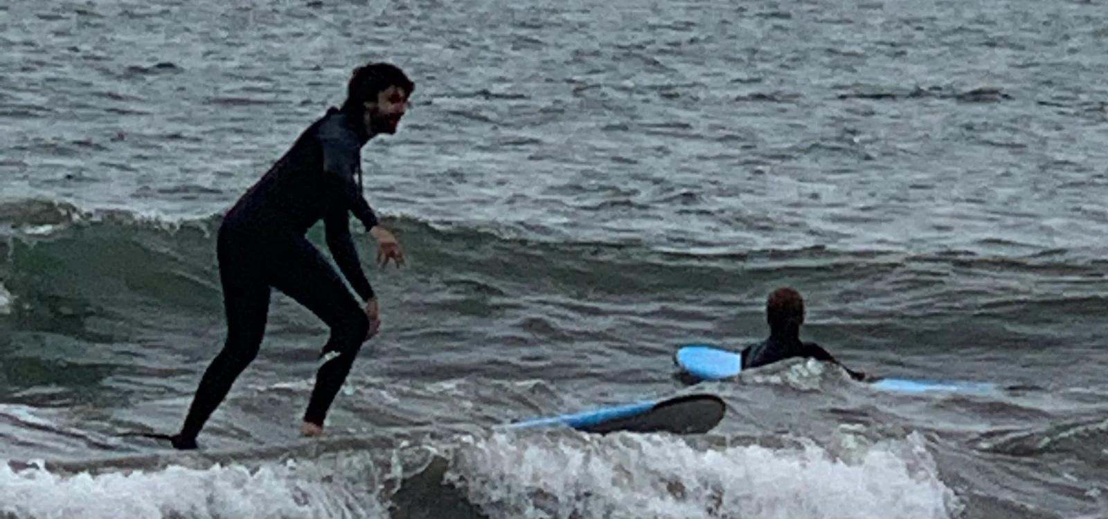 Riding a small wave in a black wetsuit, with another surfer in the water behind.